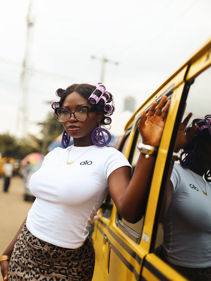 Fashionable woman with hair rollers poses by a yellow bus in a vibrant Lagos street.